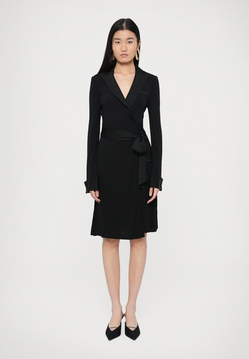 Μαύρο φόρεμα κρουαζέ με μακριά μανίκια, φαρδύ γιακά, ζώνη με φιόγκο στη μέση και υφή υφάσματος. Συνδυασμένο με μυτερές μαύρες γόβες, που αναδεικνύουν μια κομψή σιλουέτα.