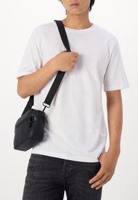 Zwarte crossbody tas van glad materiaal, met ritssluiting en verstelbare schouderband, gedragen over een wit T-shirt en donkere jeans.