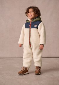 Fleecový overal v krémovej farbe s tmavozelenou hornou časťou a zelenou kapucňou. Obsahuje predný zips a nášivku s logom, skombinovaný s hnedými turistickými topánkami.