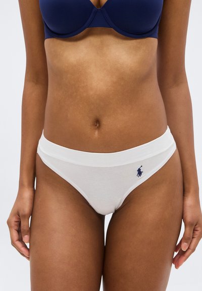 Femme portant un soutien-gorge bleu marine et un slip de style bikini blanc avec un petit logo joueur de polo bleu marine sur la hanche gauche, visible de la moitié du torse aux cuisses.