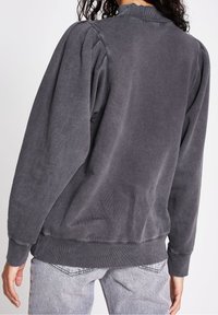 Grauer Pullover mit puffierten Ärmeln und einem gerippten Kragen. Weiche Textur,abschließende Säume und eine lockere Passform. Kombiniert mit hellgrauen Jeans.