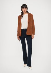 Brown suede blazer over a light sweater, paired with dark flared jeans. White pointed-toe shoes complete the outfit. Simple, tailored design.