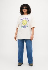 Beige oversized t-shirt featuring a colorful graphic of a smiling sun and flowers. Paired with blue straight-leg jeans and dark footwear.