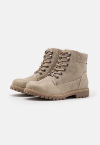 Une paire de bottines en suede beige à lacets, avec des semelles en caoutchouc marron et des œillets en métal, orientées vers la droite sur un fond blanc.