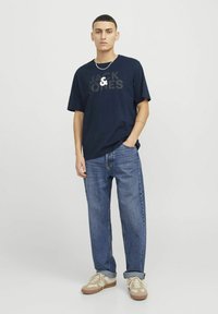 Tume sinine t-särk suure "JACK & JONES" halli logoga, millele on sobitatud lõdva lõikega sinised denimpüksid ja beežid sneakersid valgete detailidega.