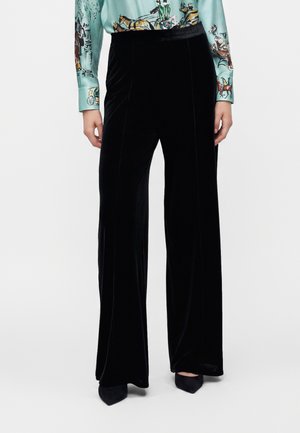 Pantalon large en velours noir porté avec des chaussures pointues noires et une chemise bleu clair avec des manches imprimées de chevaux et de carrosses.