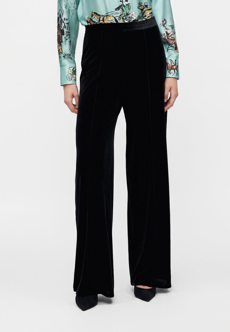 Pantalon large en velours noir porté avec des chaussures pointues noires et une chemise bleu clair avec des manches imprimées de chevaux et de carrosses.