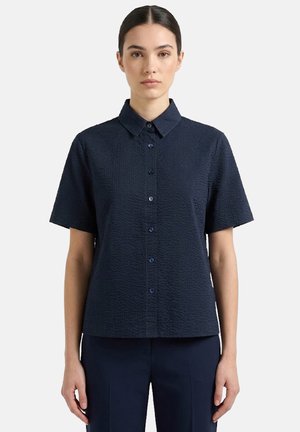 Donna che indossa una camicia blu navy a maniche corte con bottoni e tessuto strutturato, abbinata a pantaloni coordinati, in piedi davanti a uno sfondo chiaro e semplice.