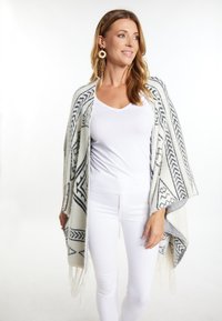 Sciarpa intrecciata di colore crema con motivi geometrici neri, caratterizzata da orli frangiati. Modella che indossa una maglietta bianca e jeans, in piedi contro uno sfondo chiaro.