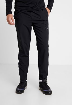 Persoon draagt zwarte Nike trainingsbroek en zwarte Nike sneakers, staat met één hand in de zak tegen een effen achtergrond.