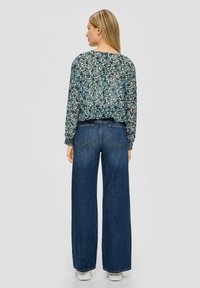 Bluse mit floralem Druck, langen Ärmeln und rundem Ausschnitt, kombiniert mit hochgeschnittenen, weiten dunkelblauen Jeans und weißen Turnschuhen.