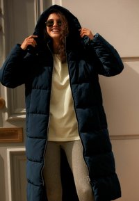 Abrigo largo acolchado azul marino con capucha, con cierre de cremallera y bolsillos laterales, llevado sobre una camisa amarilla clara y leggings grises con textura.