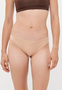 Vrouw in beige katoenen bikiniondergoed en een bruine sportbeha, staand tegen een effen witte achtergrond.