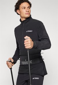 Svart fleecegenser med halv-zip, med "TERREX"-merke. Brukt sammen med en svart sele og holder turstaver med grå grep.