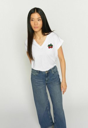 Femeie cu păr lung și negru, purtând un tricou alb cu decolteu în V și broderie cu cireșe, alături de blugi albaștri cu talie înaltă, pe un fundal simplu.