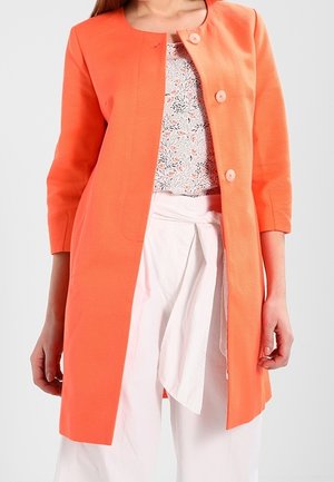 Nainen, jolla on päällään korallinoranssi pitkä takki, jossa on kolme nappia, kukkaprintillinen toppi ja valkoiset korkeavyötäröiset housut, joissa on solmittu vyö.