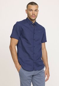 Chemise à manches courtes bleu marine à boutons avec un tissu texturé, petit col et détail de logo discret, portée avec un pantalon bleu clair.