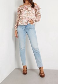 Donna che indossa un blusa floreale con spalle scoperte, jeans a vita alta blu chiaro con dettagli in perle e scarpe con tacco piatto marroni, in piedi contro un muro bianco.