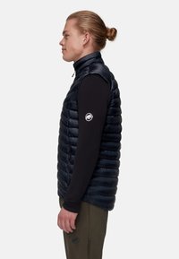 Marineblaue Steppweste mit hohem Kragen, gesteppter Optik und Reißverschluss. Getragen über einem schwarzen Langarmshirt mit einem Logo am Ärmel.