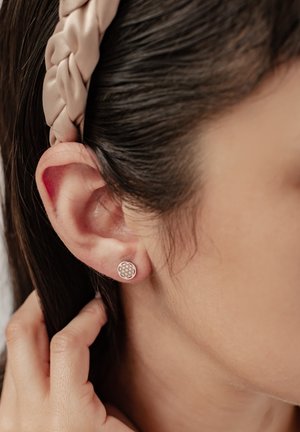 Pendiente de plata redondo con un patrón geométrico, que se lleva en el lóbulo de la oreja. El cabello está peinado con una diadema trenzada de tono claro.