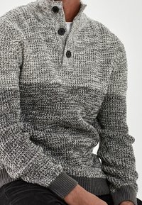 Man draagt een grijs-zwarte gebreide trui met drie zwarte knopen bij de kraag, zittend met handen op de schoot.