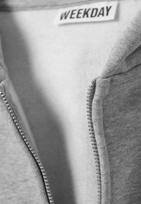 Gros plan d'un sweat à capuche gris partiellement dézippé avec une étiquette de vêtement "WEEKDAY" à l'intérieur du col.