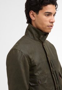 Ελιόχρωμο μπουφάν με ψηλό γιακά, φτιαγμένο από υφασμάτινο υλικό με υφή. Το μπροστινό μέρος διαθέτει πτυχώσεις και δύο τσέπες με κουμπιά. Διακριτές λεπτομέρειες ραφής.