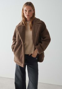 Bruine pluche fleece jas met een kraag, oversized pasvorm en voorkwasten, gelaagd over een crèmekleurige geribbelde trui en donkere jeans.