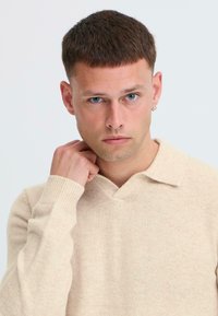 Pullover in maglia beige con scollo a V, polsini a costine e trama fine, indossato da un modello con capelli corti e orecchini.