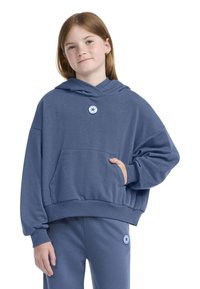 Hanorac albastru dintr-un material moale, dotat cu o buzunar frontal și un patch cu logo pe piept. Asortat cu pantaloni de trening albaștri.