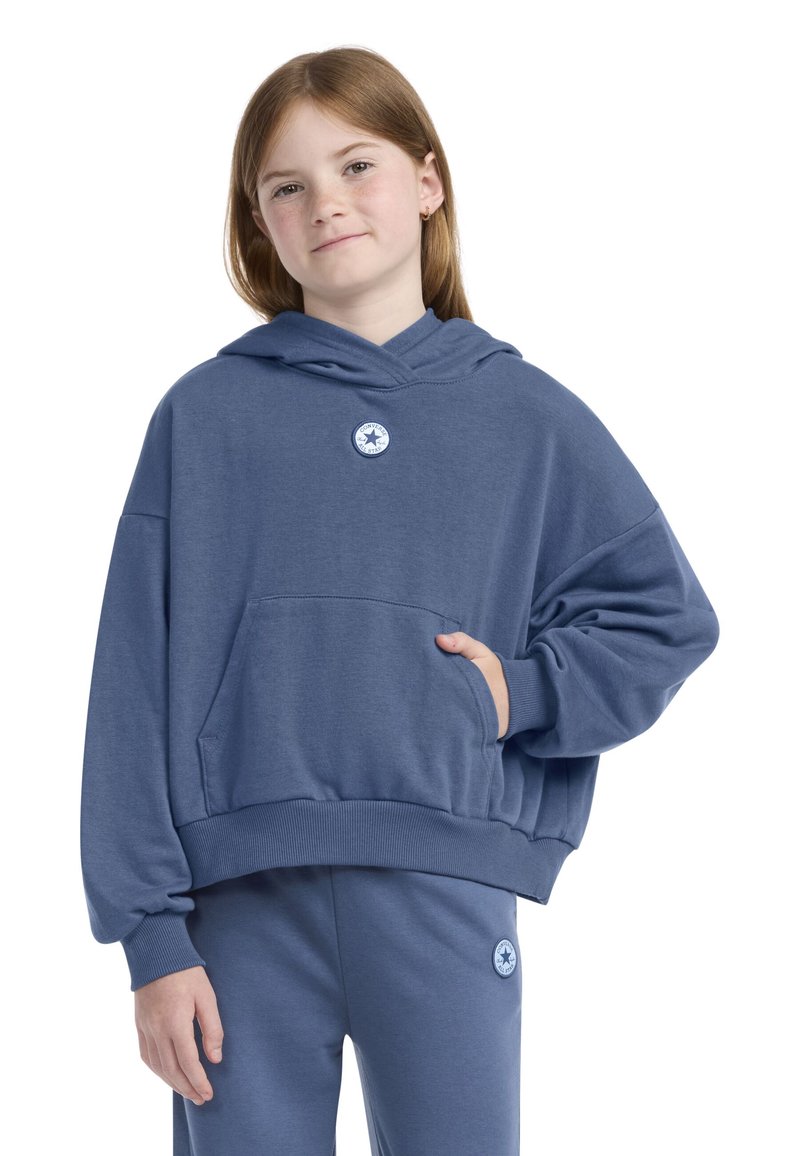 Hanorac albastru dintr-un material moale, dotat cu o buzunar frontal și un patch cu logo pe piept. Asortat cu pantaloni de trening albaștri.