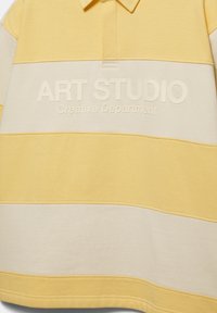 Chemise à manches longues rayée jaune et beige avec un col, affichant le texte en relief "ART STUDIO Creative Department" sur la poitrine.