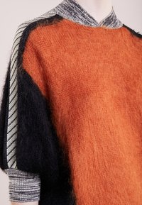 Strickpullover mit einem orangefarbenen, flauschigen Körper, schwarzen Ärmeln und einem grau gestreiften Kragen. Die strukturierte Wolle verleiht ein gemütliches Erscheinungsbild.