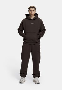 Brun hettegenser med frontlomme og logo, kombinert med matchende joggebukser med sidelommer. Stoffet ser mykt og strukturert ut.