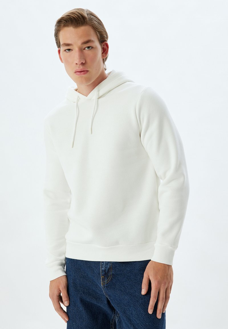 Witte hoodie van zacht materiaal, met een voorzak, verstelbare koordjes en geribbelde manchetten. Gepaarde met donkerblauwe spijkerbroek.