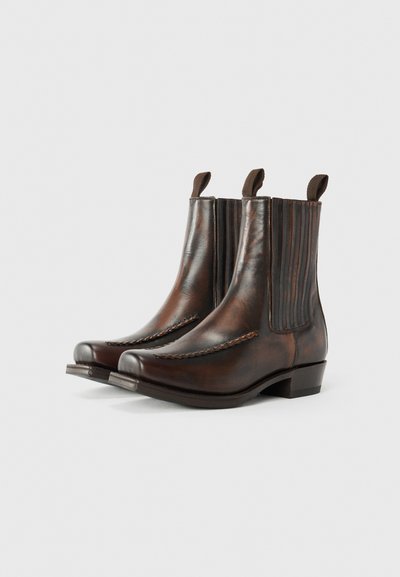 Botas tobilleras de cuero marrón con paneles laterales elásticos, costuras decorativas en la puntera y dos tiradores para facilitar su uso. El tacón plano proporciona estabilidad.
