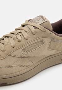 Beige mockaskor med texturerad gummisula, platta snörningar, vadderad tunga och Reebok-logga på sidan och tungan.