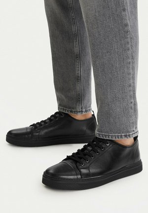 Osoba nosící černé kožené nízké tenisky s černými tkaničkami a šedé džínové kalhoty, stojící na bílém povrchu.