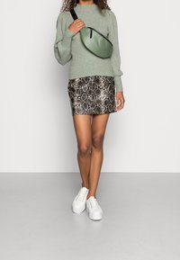 Pull tricoté vert clair, mini-jupe à motif serpent noir et beige, et baskets blanches. Le sac est d'un vert élégant avec des accents de sangle noirs.