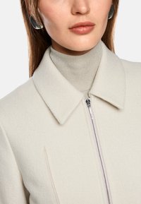 Light-colored wool coat with a collared neckline and front zipper. Styled over a textured beige turtleneck. Features large silver earrings.