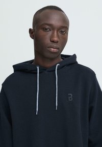 Marineblauwe hoodie met een gestructureerde afwerking, voorzien van een capuchon met trekkoorden, contrasterende lichtgrijze veters en een subtiel zwart logo op de borst.