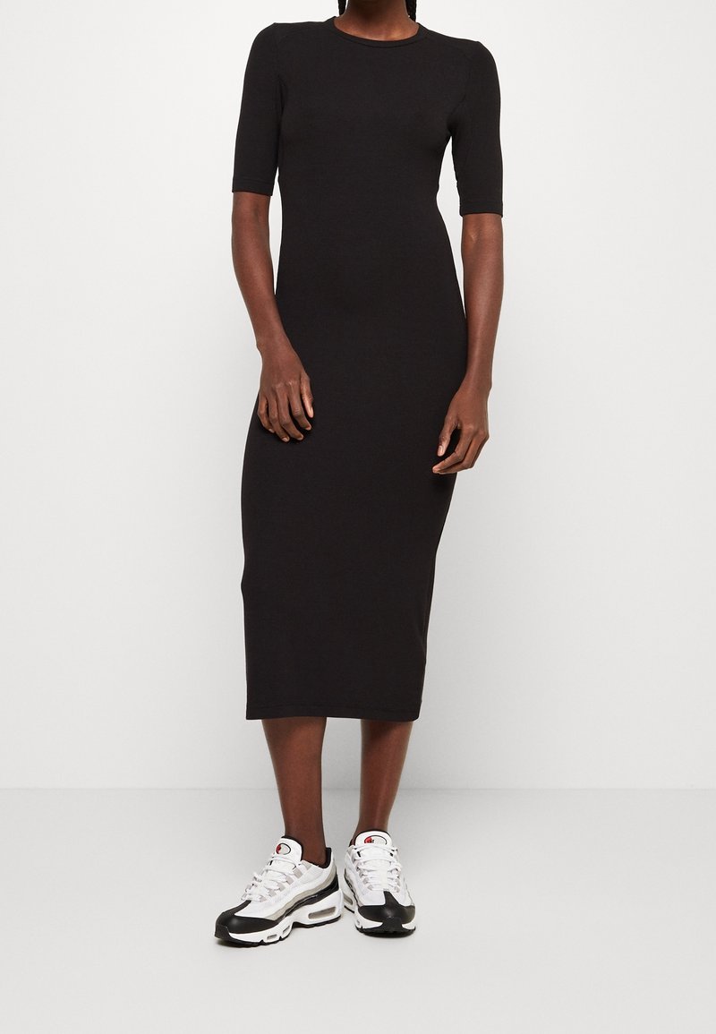Robe noire midi à manches courtes, fabriquée à partir d'un tissu extensible. Associée à des baskets blanches et noires, se tenant contre un fond uni.