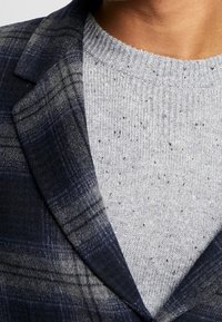 Un blazer a quadri navy si sovrappone a un maglione a maglia leggera grigio chiaro e maculato. Il blazer presenta un colletto a punta e un tessuto testurizzato.