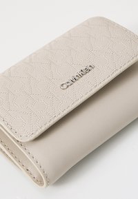 Calvin Klein kreminis piniginė, pagaminta iš tekstūruotos sintetinės medžiagos, su geometriniu reljefiniu raštu ir sidabriniu logotipu ant priekinės dalies.