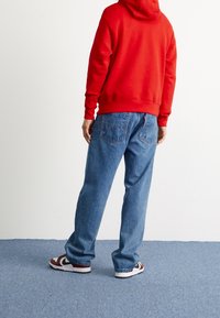 Röd huvtröja, blå denimjeans och vinröda/vita sneakers. Jeansen har stora bakfickor och en avslappnad passform.