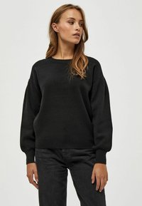 Črn pleten pulover z ohlapnim krojem, rebrastimi manšeti in zaobljenim izrezom, v kombinaciji s temnimi jeans hlačami. Mehka tekstura in preprost dizajn.