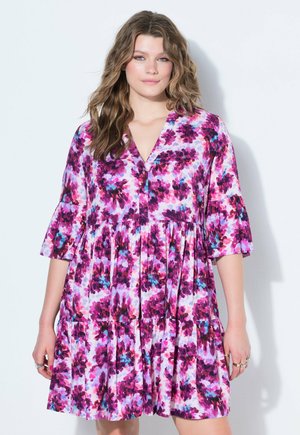 Femme portant une robe fleurie longueur genou avec des motifs roses, violets et bleus et des manches trois-quarts, debout sur un fond blanc.