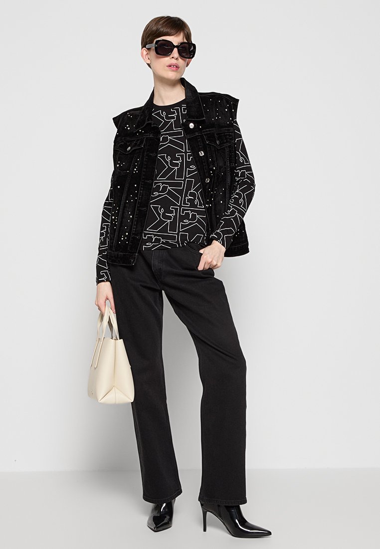 Gilet in denim nero con borchie argento, camicia a maniche lunghe con motivo, pantaloni neri, tacchi a punta in vernice e borsa a spalla color crema.