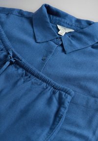 Cămașă albastră din bumbac, cu guler și nasturi pe partea din față, împreună cu pantaloni cu talie elastică asortată, așezate pe o suprafață deschisă la culoare.