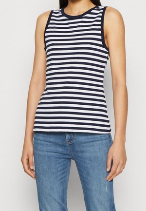 Mujer vistiendo una camiseta sin mangas de rayas horizontales negras y blancas combinada con jeans azules de mezclilla, de pie contra un fondo liso.